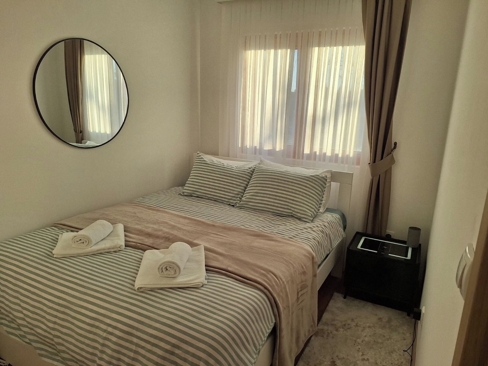Een slaapkamer met een bed, een spiegel, een raam met gordijnen en een nachtkastje met een lamp.