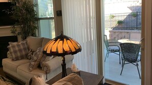 Una sala de estar con sofá, lámpara, planta y vista a un patio exterior con mesa y sillas.