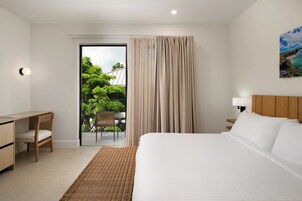 Habitación de hotel con cama, escritorio, silla, balcón con vista a vegetación y un cuadro en la pared.