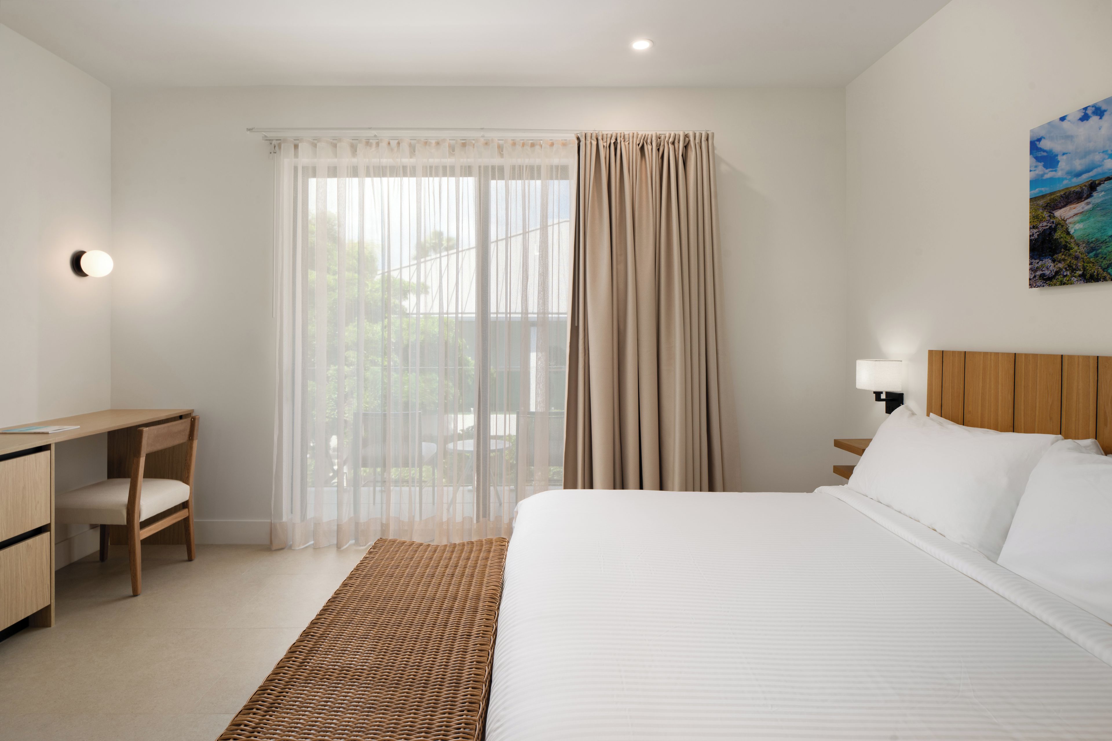 Quarto de hotel com cama, escrivaninha, cadeira, luminária e um quadro na parede.