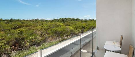 Ein Balkon mit Glastrennwand, der auf eine grüne Landschaft blickt. Zwei weiße Stühle und ein kleiner runder Tisch mit einem Weinglas stehen bereit.
