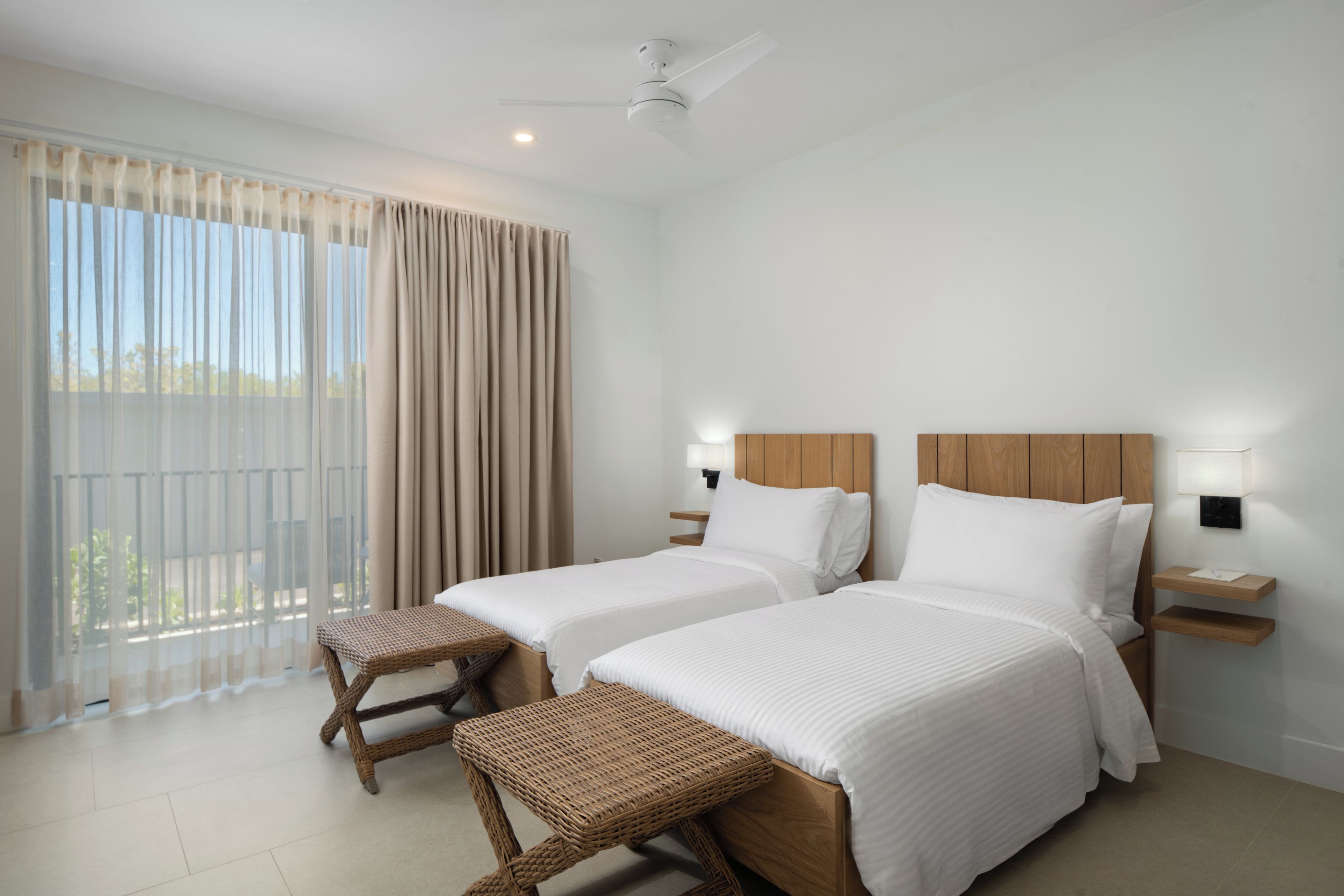 Habitación de hotel con dos camas, ventilador de techo, ventana grande con cortinas y balcón.