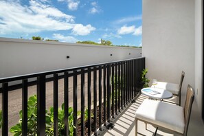 A balcony with a white table and chairs, a black metal railing, and a view of the sky and greenery.