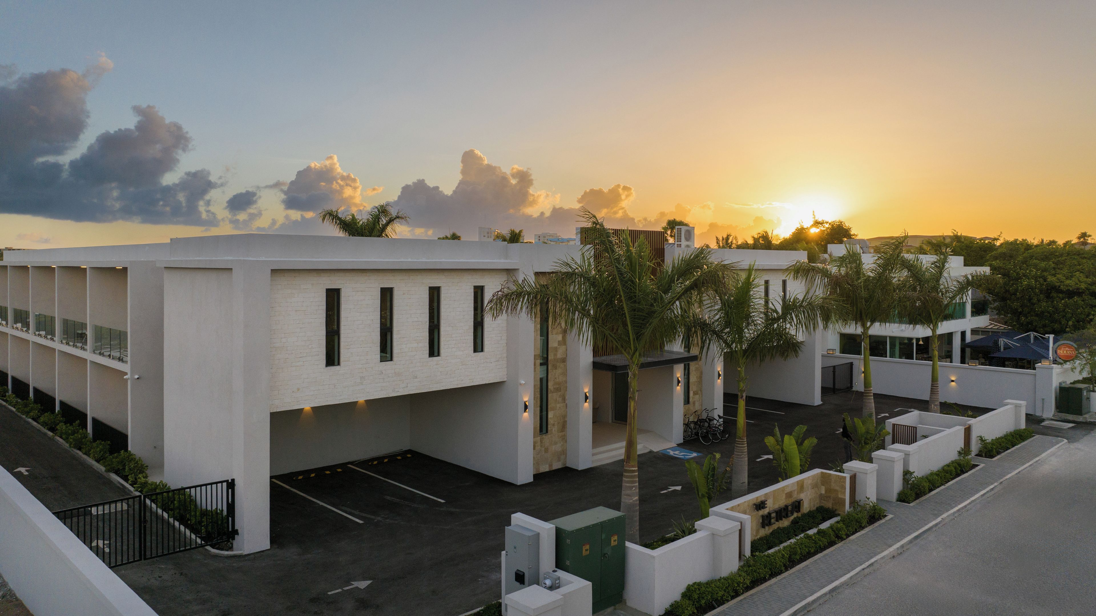 Un edificio moderno con estacionamiento, palmeras y una puesta de sol al fondo.