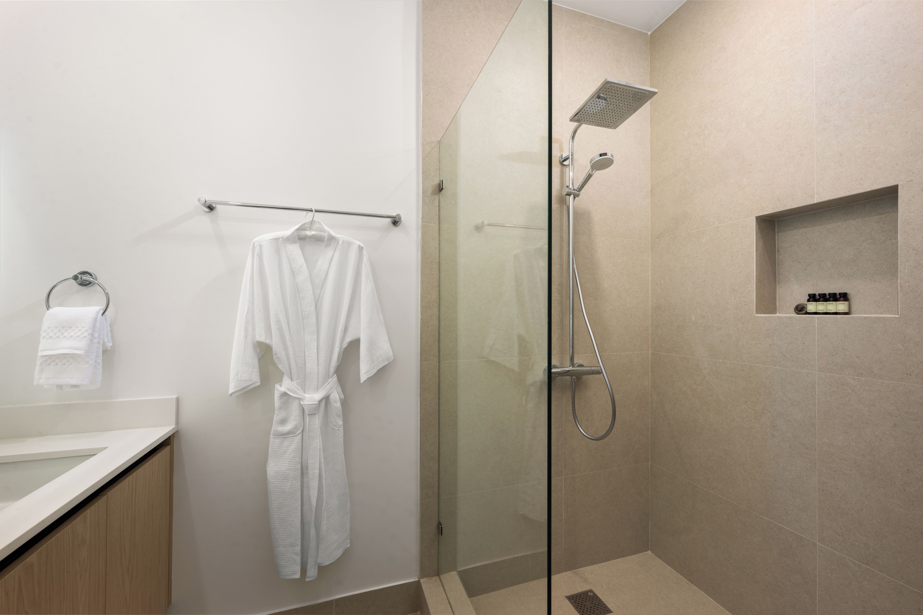 Un baño moderno con ducha de vidrio, una bata blanca colgada en un perchero y un estante pequeño con artículos de tocador.