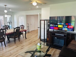 Ein Wohnzimmer mit Fernseher, der verschiedene Streaming-Dienste anzeigt, einem Glastisch mit Fernbedienung, einem Kamin und einem Essbereich mit Holzsesseln und einem Tisch.
