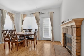 A dining room with a wooden table and chairs, a fireplace, and a window with curtains.