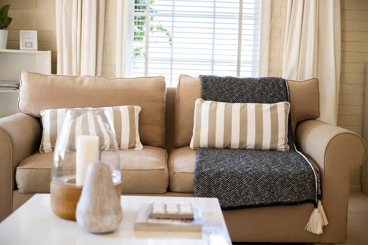 A beige sofa with striped and polka-dotted cushions, a glass candle holder, and a small book on a white table.