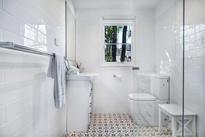 Ein kleines Badezimmer mit weißen Fliesen, einem Fenster, einer Toilette und einer kleinen Sitzbank.