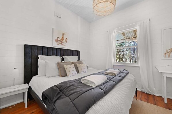 Um quarto com uma cama grande, paredes brancas, cabeceira de madeira, mesa de cabeceira, janela com cortinas e luminária pendente.