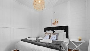 Quarto com cama, paredes brancas, piso de madeira, mesinha de cabeceira com um vaso e luminária suspensa.