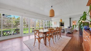 Un vaste espace dédié à la salle à manger, avec une grande table et des chaises, un luminaire suspendu en fibres tressées et de grandes fenêtres donnant sur un jardin.