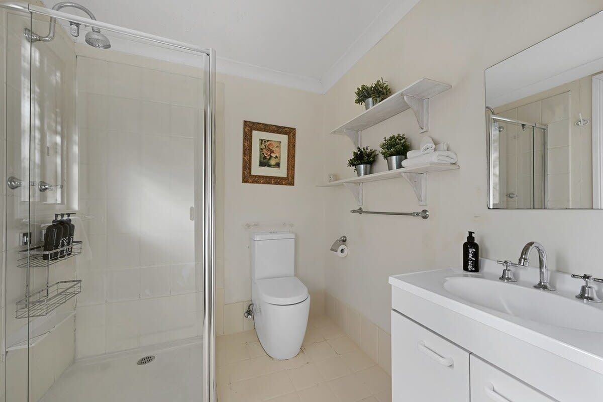 A bathroom with a glass shower enclosure, a toilet, a sink with a mirror, and shelves with toiletries.
