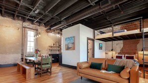 A spacious living area with a wooden floor, a sofa, a dining table with chairs, and a kitchen in the background.