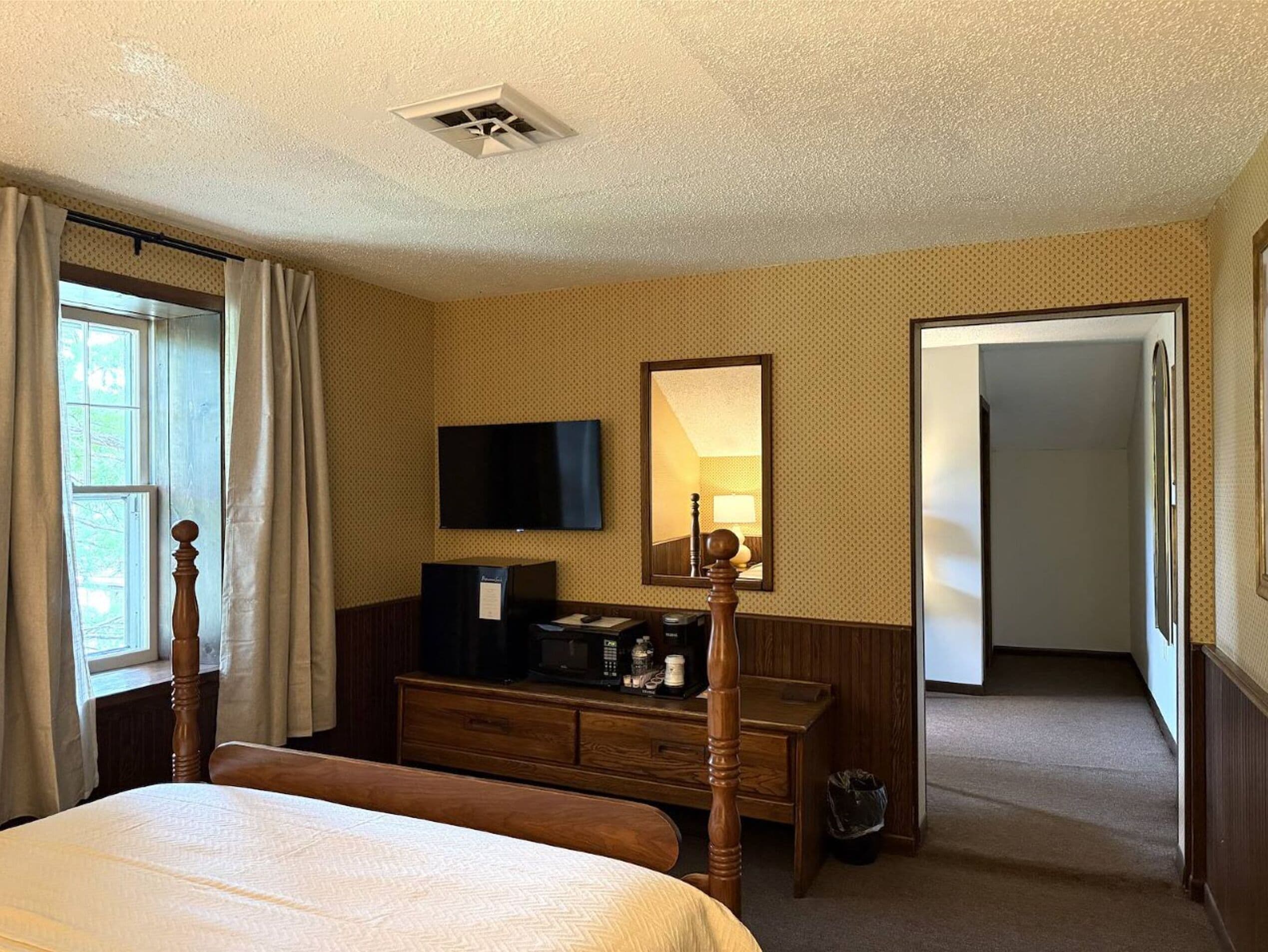Quarto de hotel com cama, cômoda, televisão, espelho e porta que dá para outro espaço.