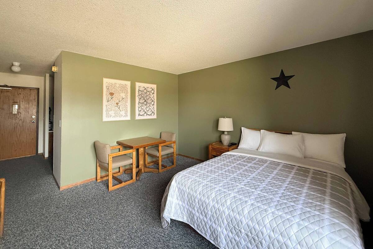 A hotel room with a bed, a desk with two chairs, a nightstand with a lamp, and two wall art pieces.
