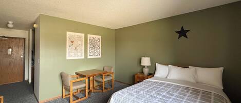 Una habitación de hotel con una cama, un escritorio con dos sillas, una mesita de noche con una lámpara y dos obras de arte en la pared.