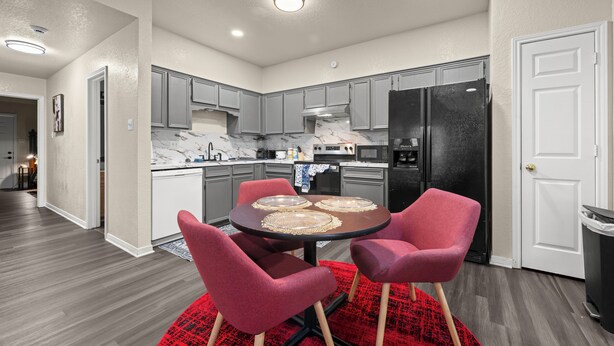 A modern kitchen with grey cabinets, a black refrigerator, and a dining area with a red rug. - Urban Oasis (Killeen)