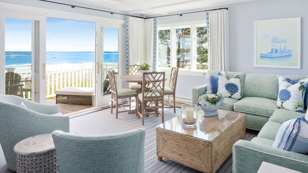 Een ruime woonkamer met uitzicht op het strand, een eethoek met houten stoelen en een groene bank met decoratieve kussens.