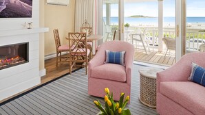 A room with a fireplace, a television, a balcony with a view of the beach, and a dining area with chairs and a table.