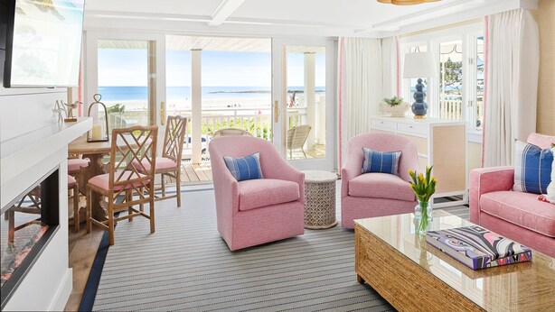 A spacious living room with a large window overlooking a beach, furnished with pink chairs, a coffee table, and a TV.