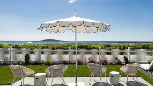 An outdoor patio with a white umbrella, wicker chairs, and a view of the sea.