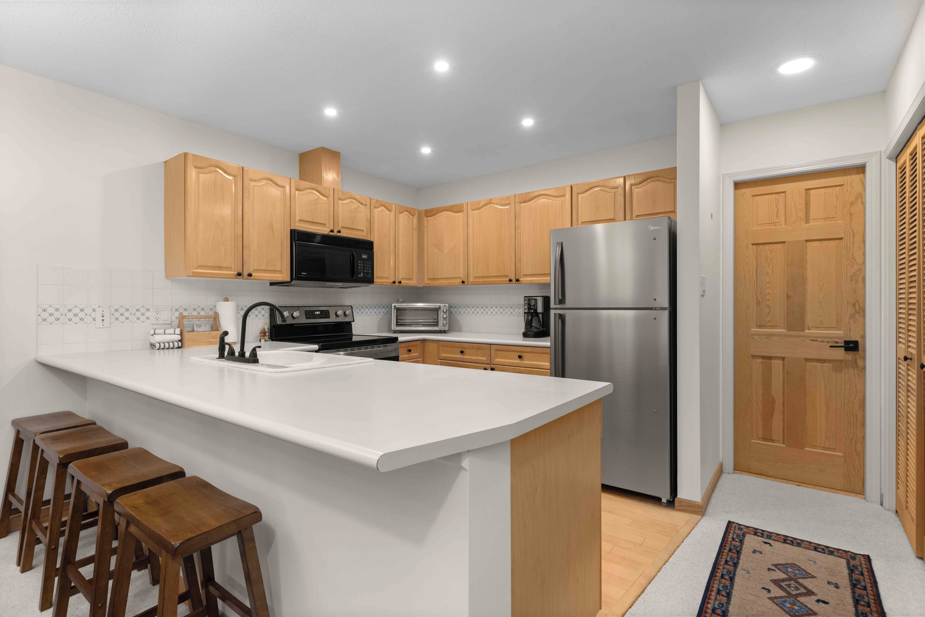 A modern kitchen with wooden cabinets, a stainless steel refrigerator, a microwave, and a sink. There are three wooden stools at the island.