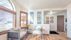 A living room with a large window, wooden flooring, a sofa, a chair, a small table with a vase, and a door leading to another room.