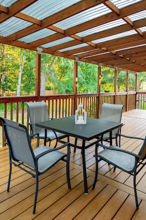 Una terraza techada al aire libre con una mesa y sillas, rodeada por una baranda de madera, con árboles de fondo.