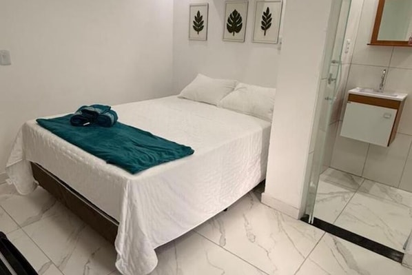 A neatly made bed with a white bedspread and a teal blanket, a small bedside table with a lamp, and a bathroom with a glass shower enclosure. - LOFTS ILHA GRANDE (Angra dos Reis)