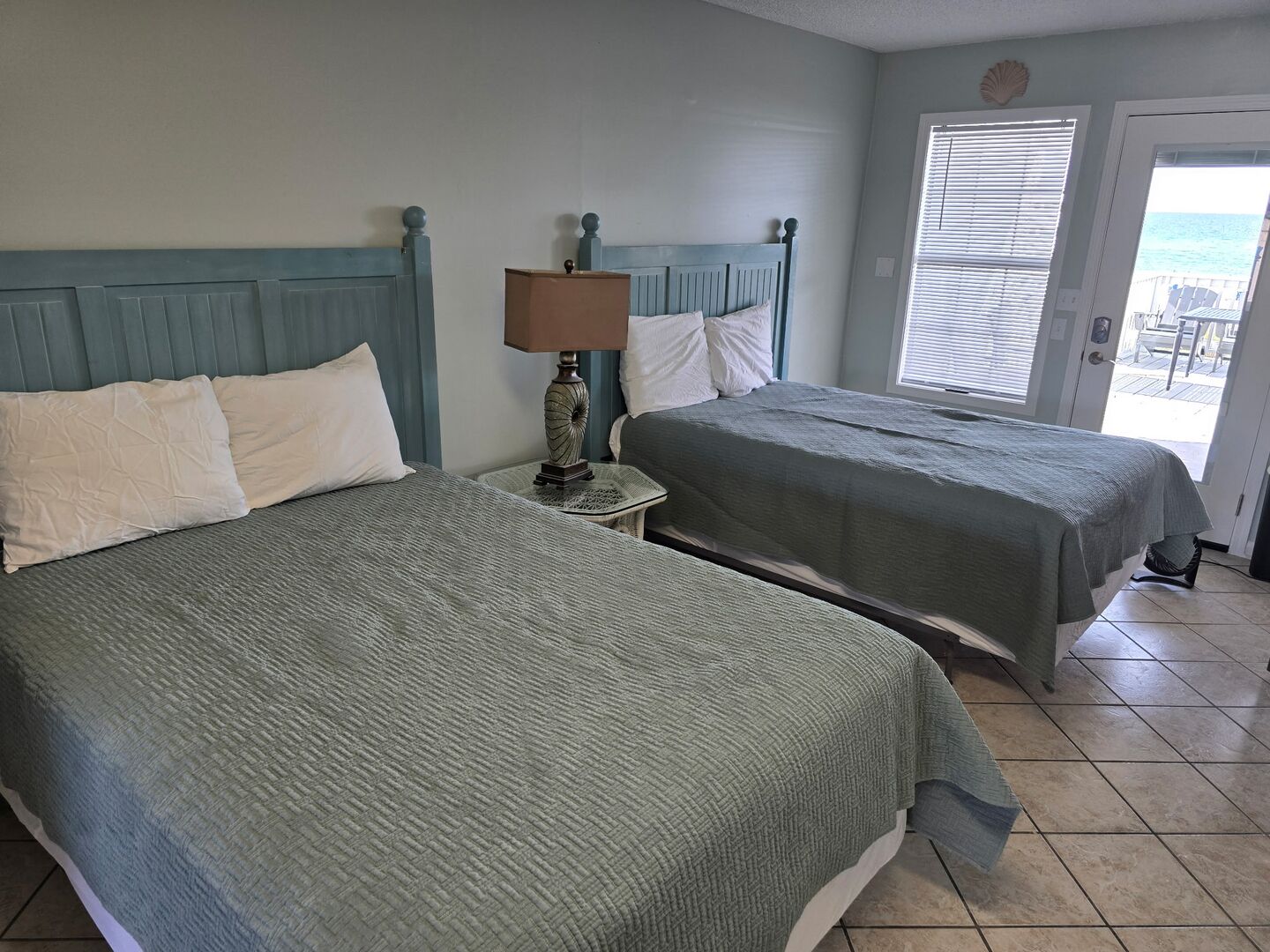 A hotel room with two beds, a nightstand with a lamp, a window with blinds, and a view of the ocean.