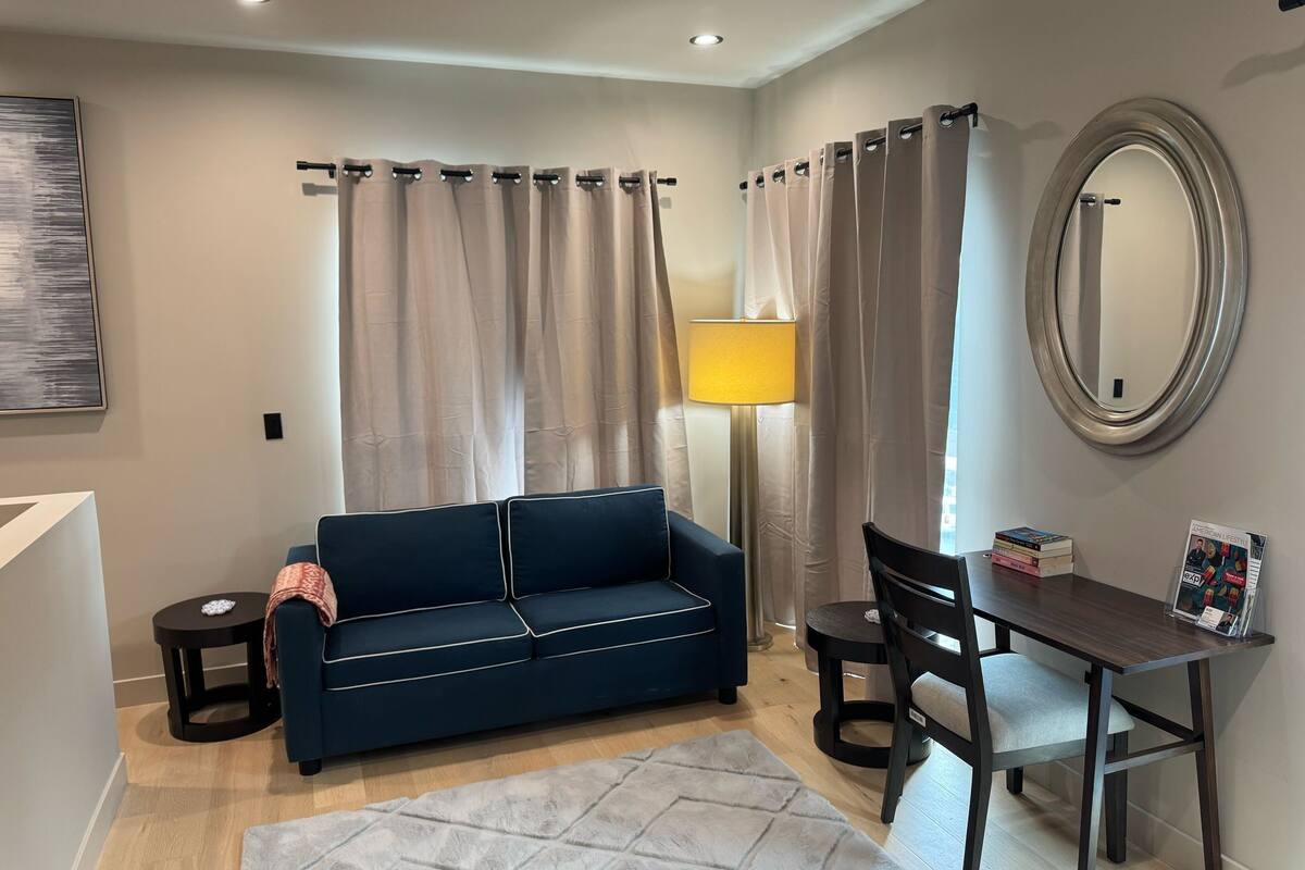 Una sala moderna con un sofá azul oscuro, una mesita redonda pequeña, un escritorio con una silla, una lámpara y un espejo grande.