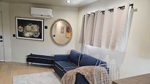 Een moderne woonkamer met een blauwe bank, een ronde spiegel, een witte airconditioner en een raam met beige gordijnen.