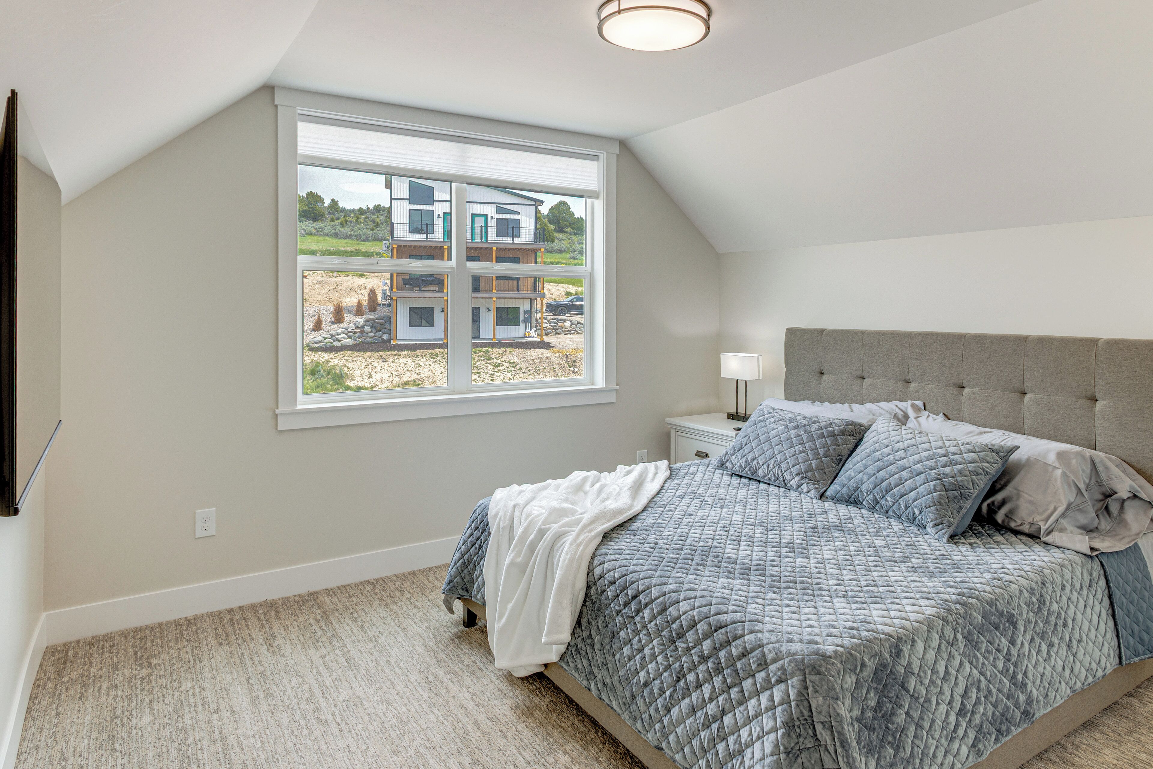 A bedroom with a large window, a bed with a grey quilt, a bedside table with a lamp, and a view of a building outside.
