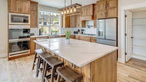 A modern kitchen with wooden cabinets, a large island with stools, stainless steel appliances, and a view of the outdoors.