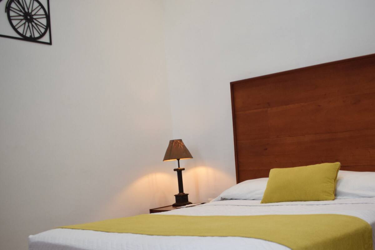 Un lit bien fait, avec une couverture jaune, une tête de lit en bois et une petite table de chevet surmontée d’une lampe.