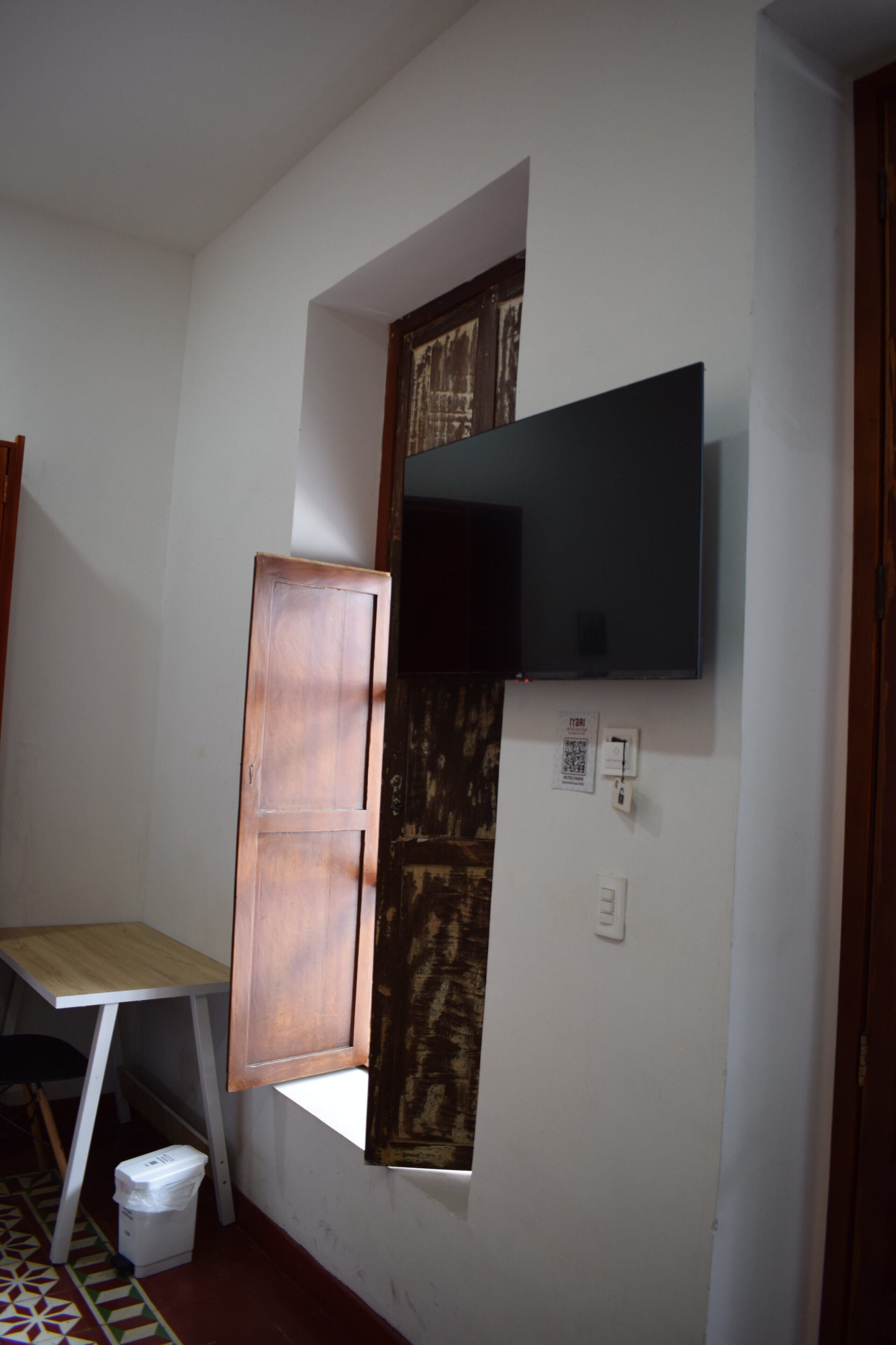 Une pièce comprenant un bureau, un téléviseur fixé au mur, une fenêtre avec un cadre en bois et une poubelle.