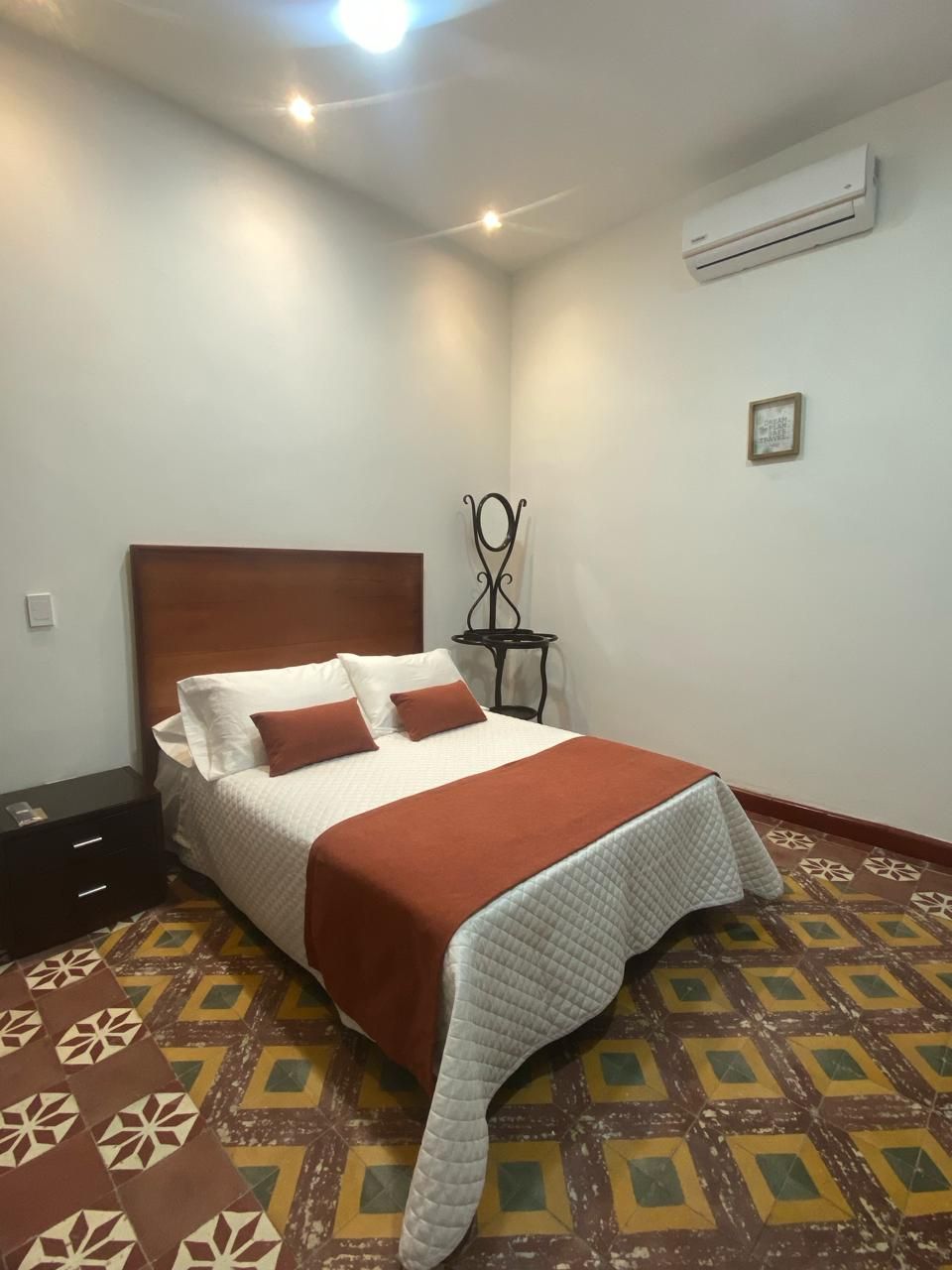 A hotel room with a bed, a nightstand, a wall-mounted air conditioner, and a patterned floor.