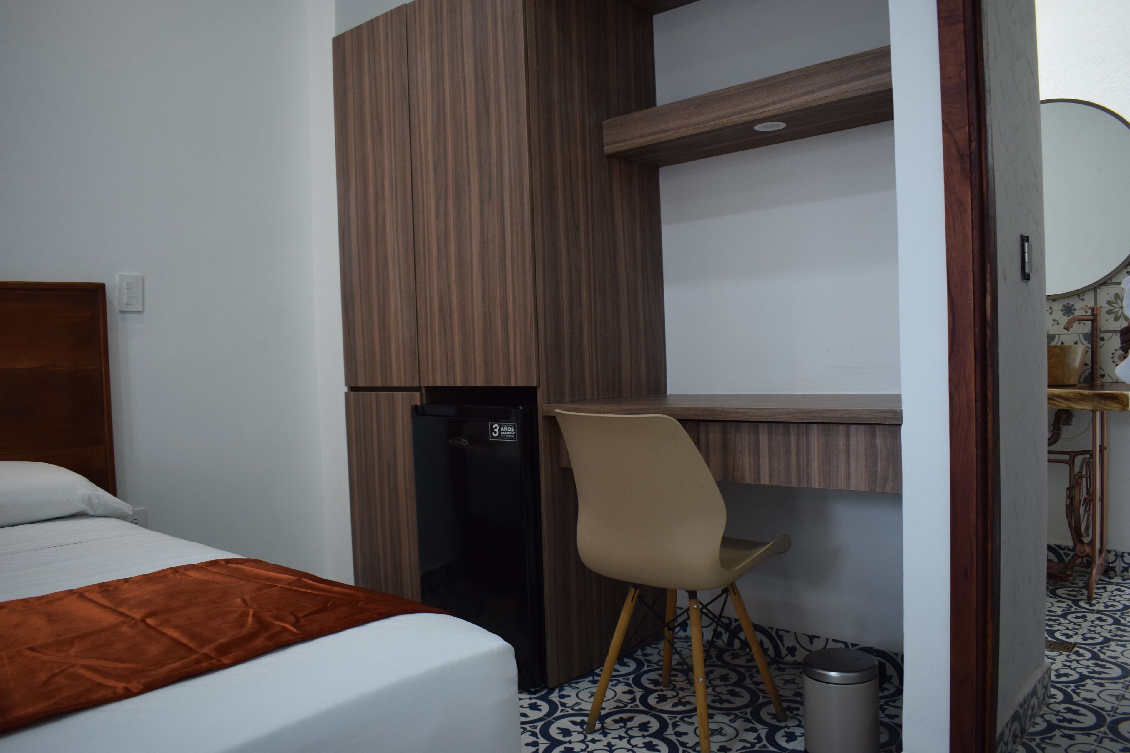 Una habitación de hotel con cama, escritorio, silla, refrigerador y basurero.