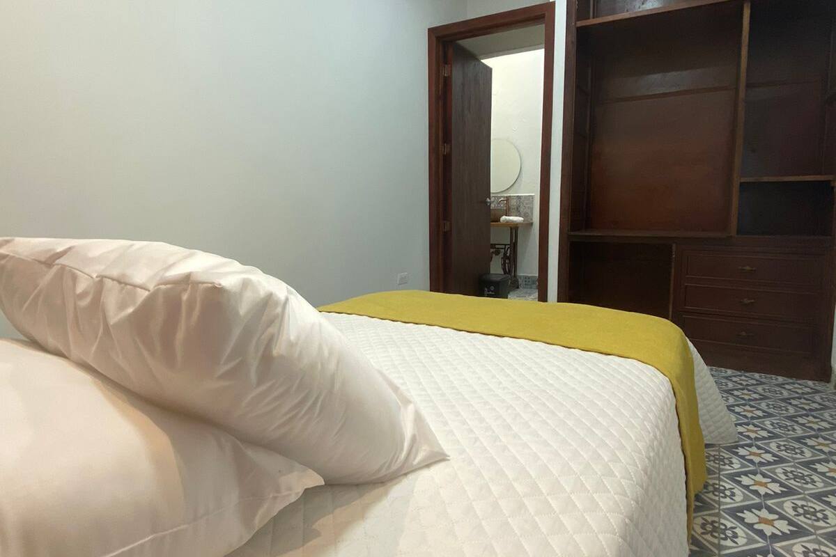 Une chambre à coucher avec un lit, une guitare, un climatiseur, une armoire et une salle de bain visible à travers une porte ouverte.