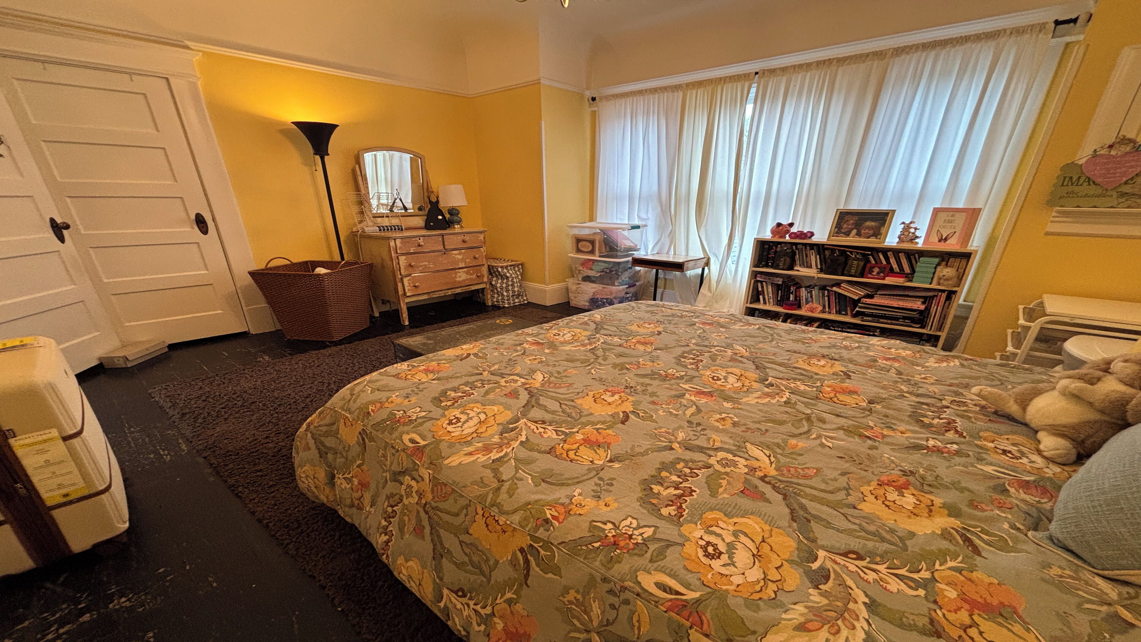 A bedroom with a floral bedspread, a dresser, a bookshelf, and a window with curtains.