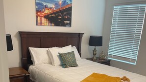 A neatly made bed with white linens and a mustard-colored throw blanket, a wooden headboard, a nightstand with a lamp, and a painting of a bridge on the wall. - Breeze Retreat (St. Augustine)