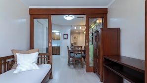 A bedroom with a bed, a wooden bench, and a desk. There is a dining area with chairs and a table in the background.