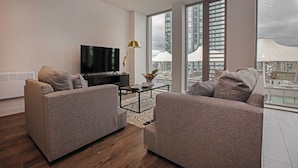 Een moderne woonkamer met twee grijze banken, een zwarte televisie, een salontafel en een vloerlamp. Grote ramen bieden uitzicht op de skyline.