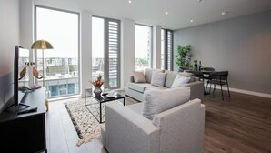 Een moderne woonkamer met een grijze bank, een salontafel en een televisie. Er is een eethoek met een tafel en stoelen. Grote ramen bieden uitzicht op de stad.