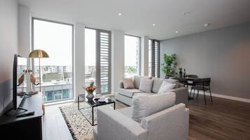 A modern living room with a grey sofa, a coffee table, and a TV. There is a dining area with a table and chairs. Large windows provide a view of the city.