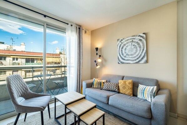 A modern living room with a grey sofa, a quilted armchair, and a small wooden table. A large window offers a view of the cityscape. - Appartement Olivier - Welkeys (Antibes)