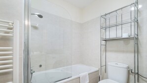 A modern bathroom with a glass-enclosed shower, a white bathtub, and a toilet with a metal shelf above it. - Appartement Olivier - Welkeys (Antibes)