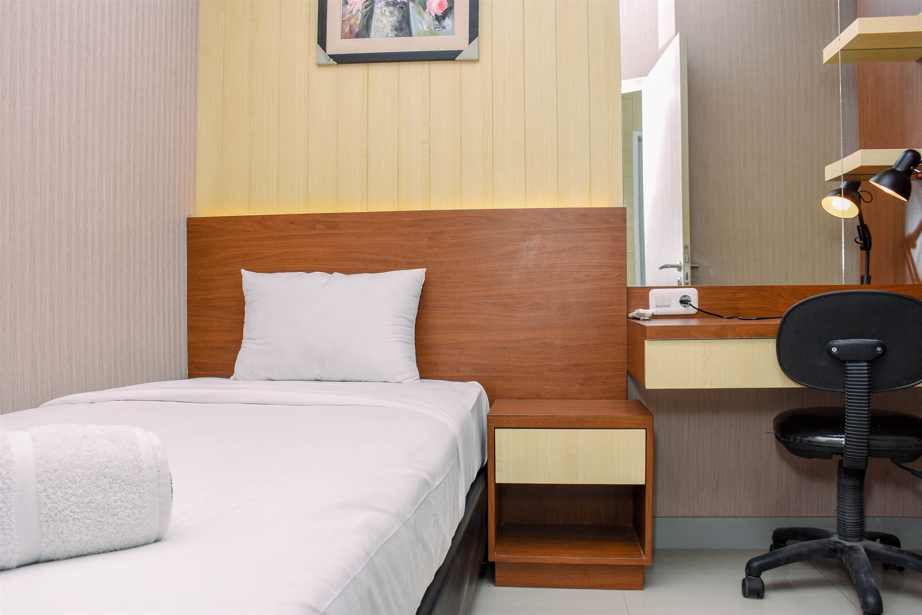 A hotel room with a wooden headboard, a bed with white bedding, a bedside table with a lamp, a desk with a chair, and a mirror.