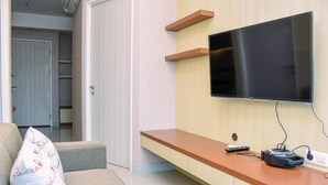 Uma sala de estar com televisão de tela plana na parede, mesa de centro de madeira, sofá com almofada decorativa e um controle remoto sobre a mesa.
